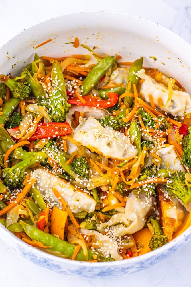 Colorful vegetable stir-fry with dumplings and sesame seeds in a white bowl.
