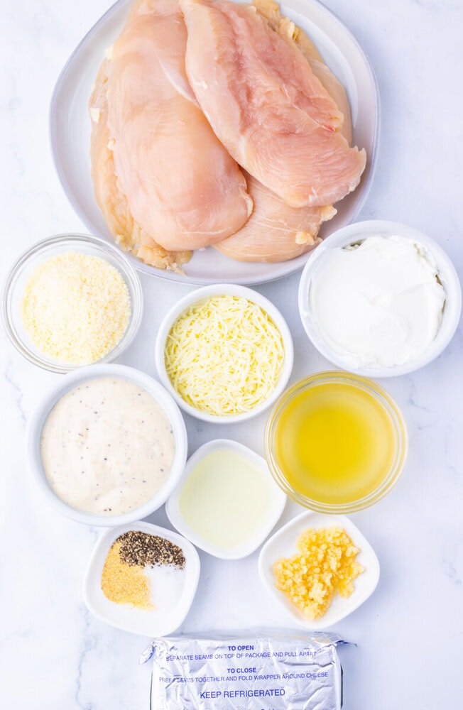 Creamy Chicken Caesar Bake ingredients laid out including raw chicken breasts, parmesan, shredded cheese, sour cream, mayo, olive oil, garlic, and corn