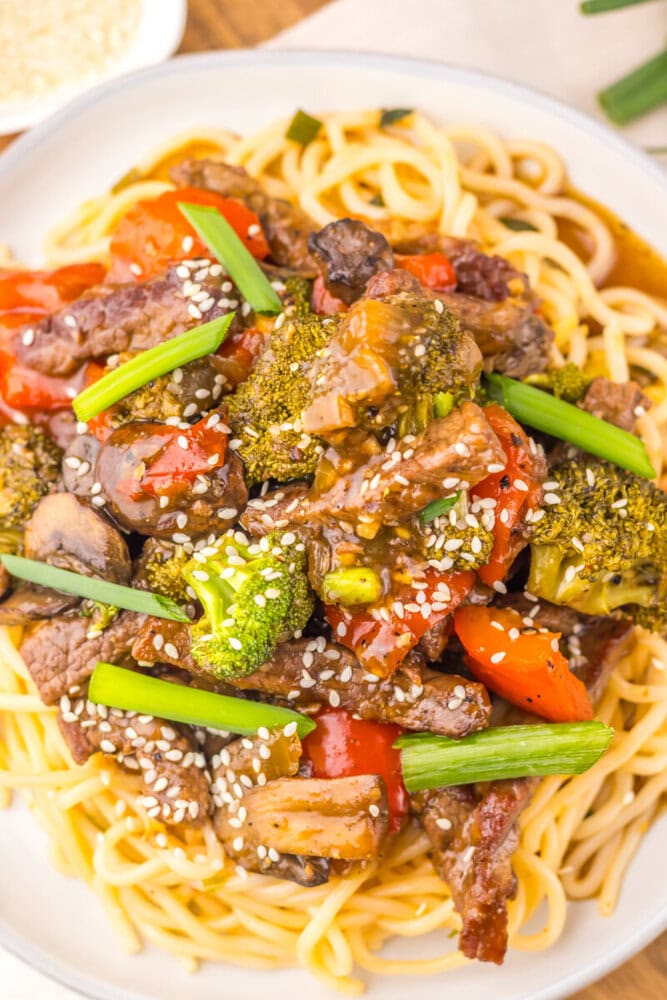 Delicious beef and vegetable stir-fry served over noodles, garnished with sesame seeds and green oni.