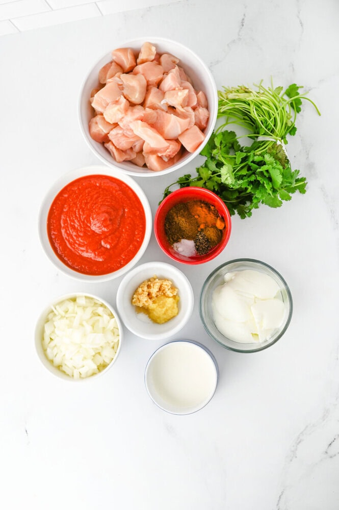 Raw chicken pieces with tomato sauce, garlic, onion, cilantro, and spices on a white surface.