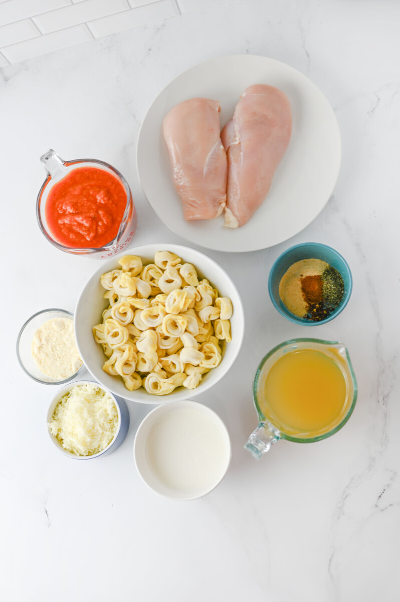 Crock pot chicken tortellini ingredients laid out including raw chicken breasts, tortellini pasta, marinara sauce, cheese, and seasonings on marble countertop