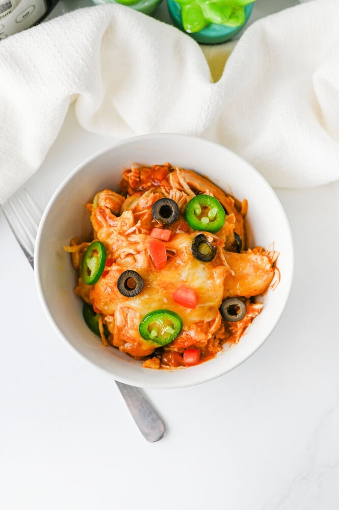 Crockpot enchiladas in white bowl topped with jalapeños, black olives, and diced red peppers
