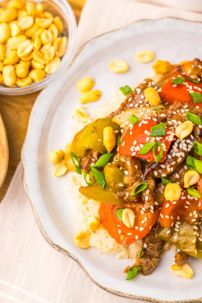 Slow cooker kung pao beef with sesame seeds, green onions, and peanuts served over rice on a white plate