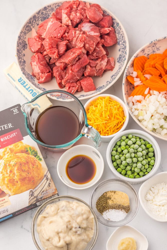 Slow cooker cheddar bay beef pot pie ingredients laid out including raw beef, vegetables, broth, and seasonings in separate bowls